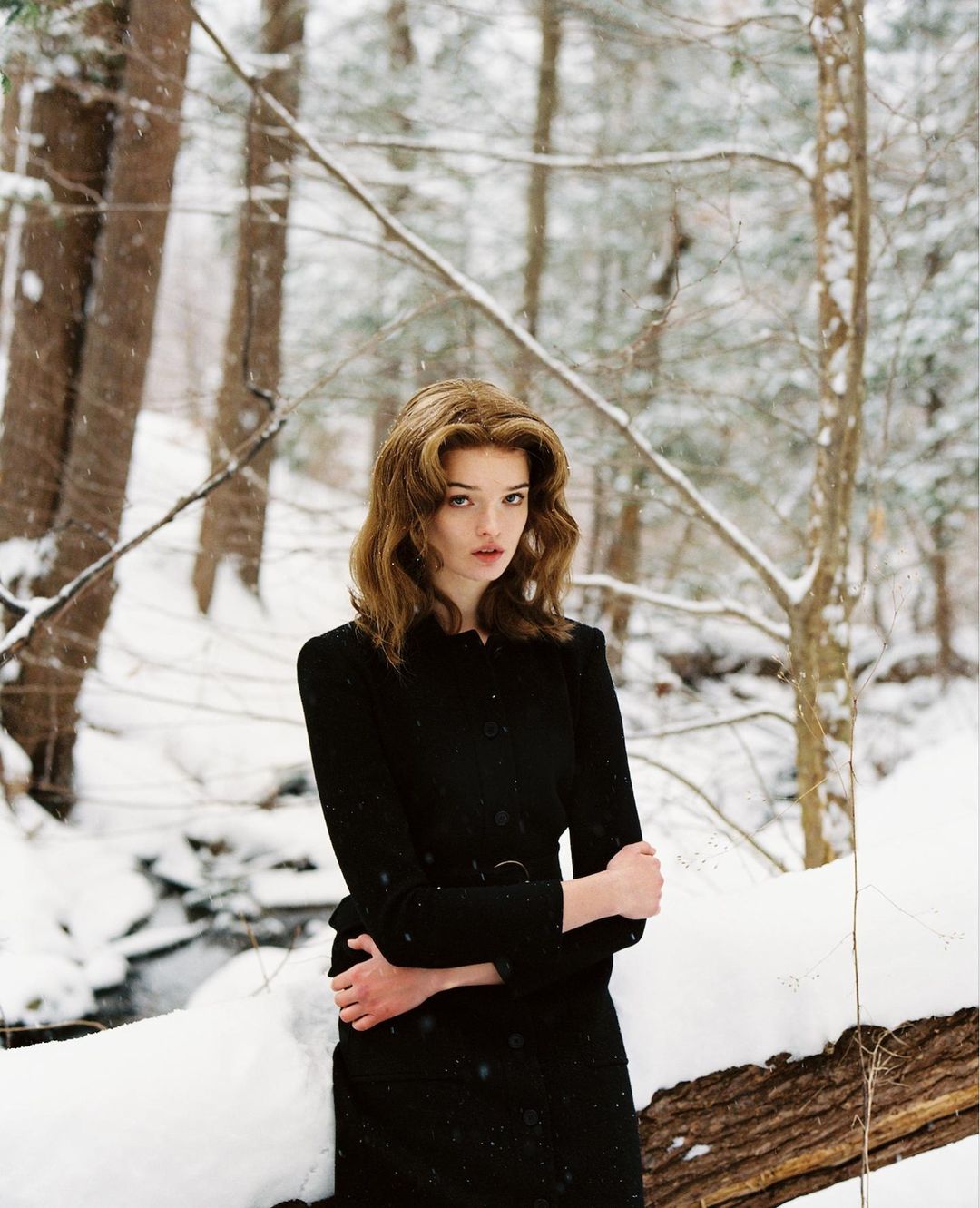 Woman in black coat stands in snowy forest. Redhead woman posing in winter landscape with bare trees, snow-covered ground, and dark coat.