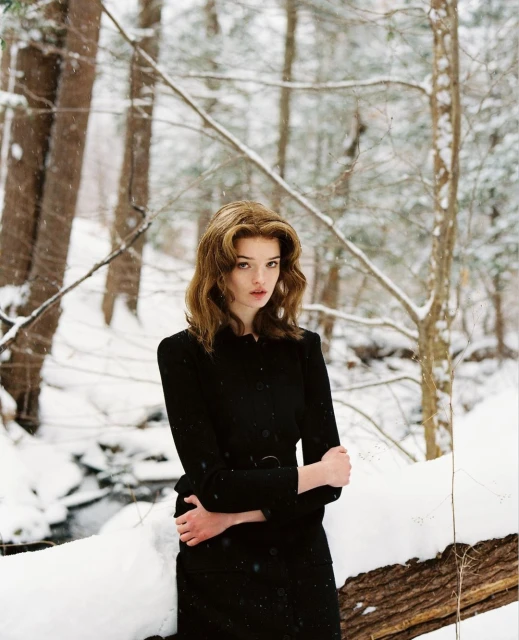 Woman in black coat stands in snowy forest. Redhead woman posing in winter landscape with bare trees, snow-covered ground, and dark coat.