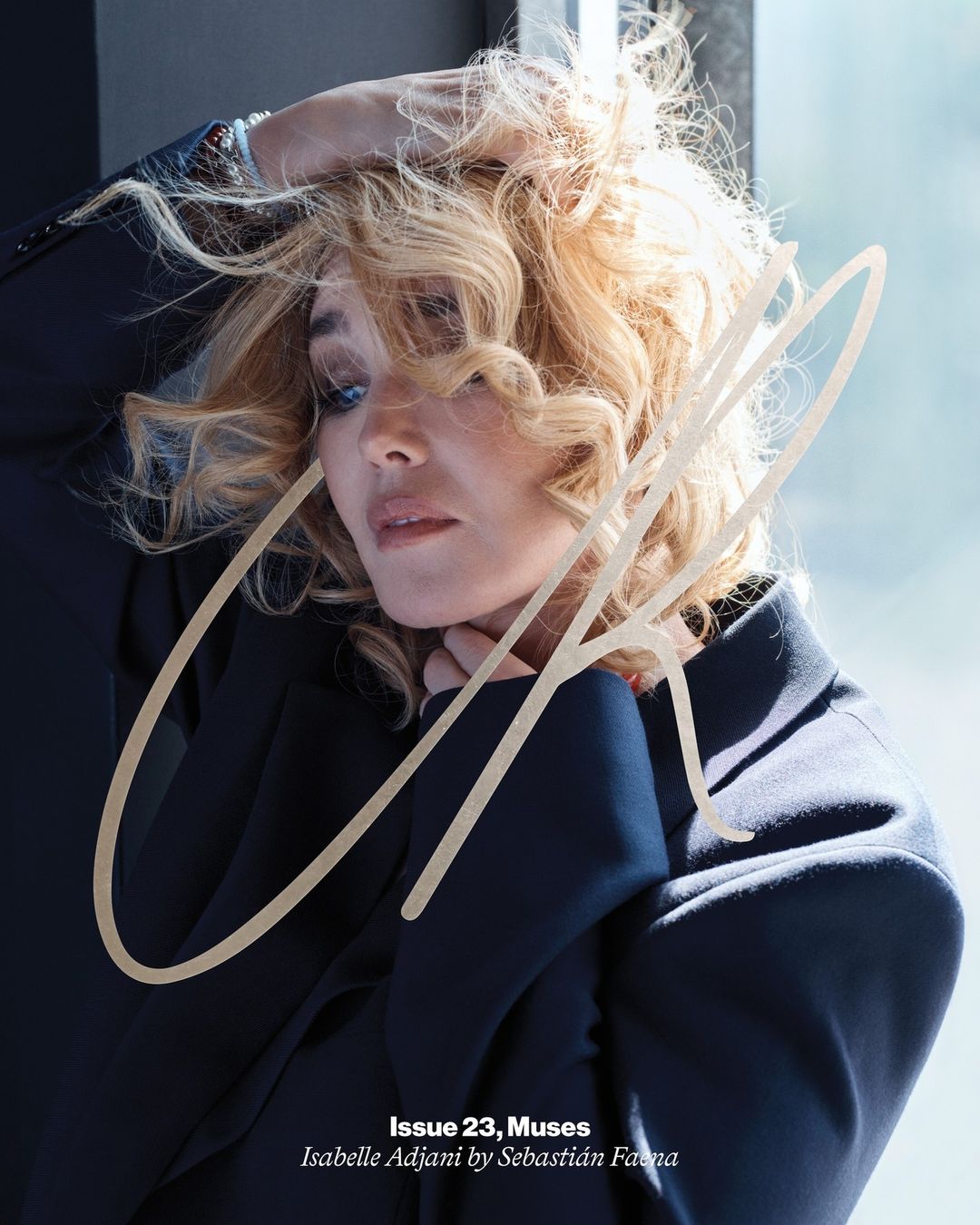 Blonde woman with curly hair wearing a dark blazer holding her hair in a portrait by Sebastian Faena