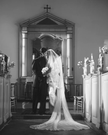 Black and white image of a bride and groom standing in a church with a long veil and bouquet of flowers on their wedding day.