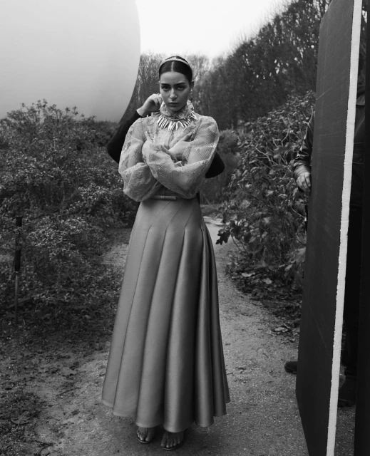 A woman stands in a garden setting, wearing a long skirt and jewelry. A person adjusts her collar. The overall image is black and white.