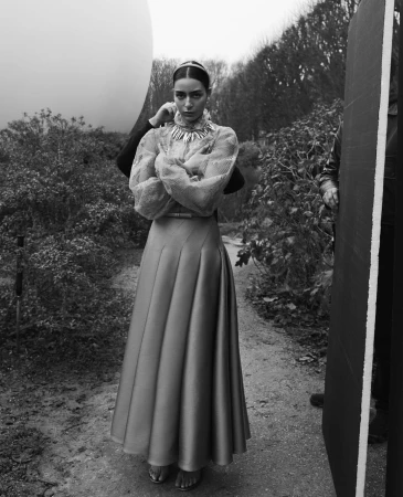 A woman stands in a garden setting, wearing a long skirt and jewelry. A person adjusts her collar. The overall image is black and white.