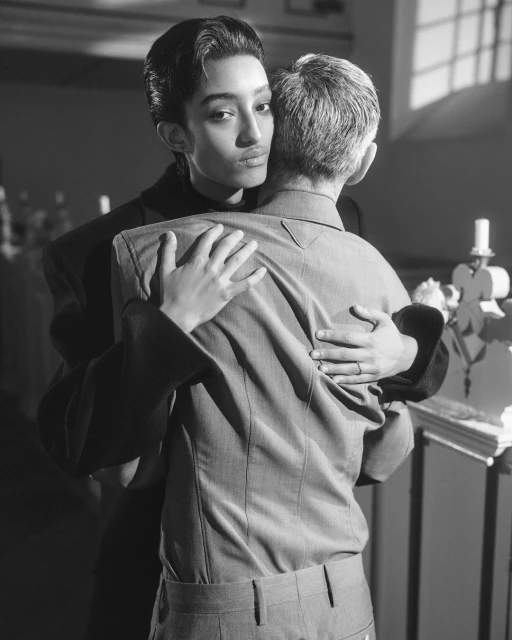 Black and white photograph of two adults embracing, one with short dark hair facing forward, the other facing away in a light colored suit.