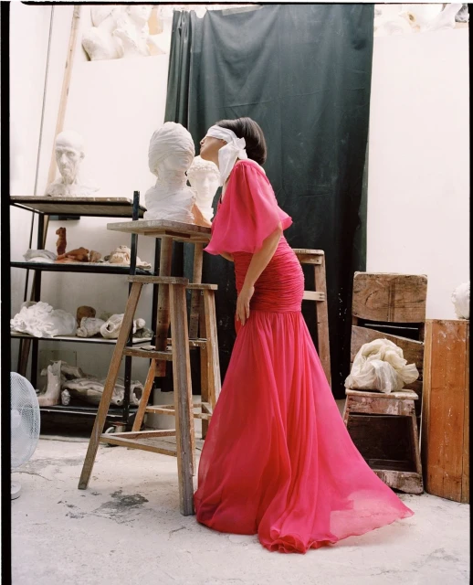 Woman in pink gown blindfolded next to sculpture in art studio with artwork on shelves and draped backdrop, creating artistic scene.