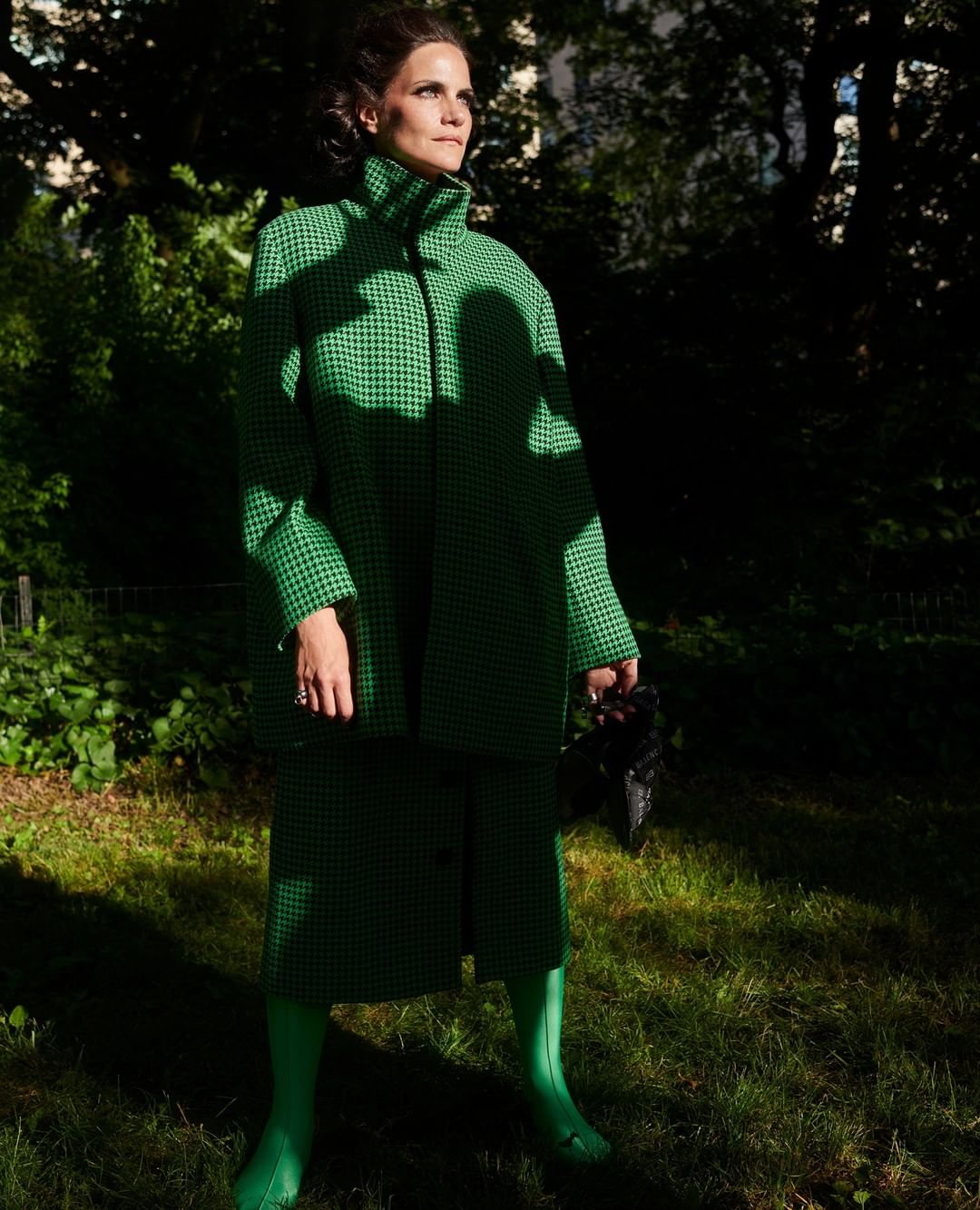 A woman in a green patterned coat and skirt standing in a grassy area, captured in natural light with a shallow depth of field.