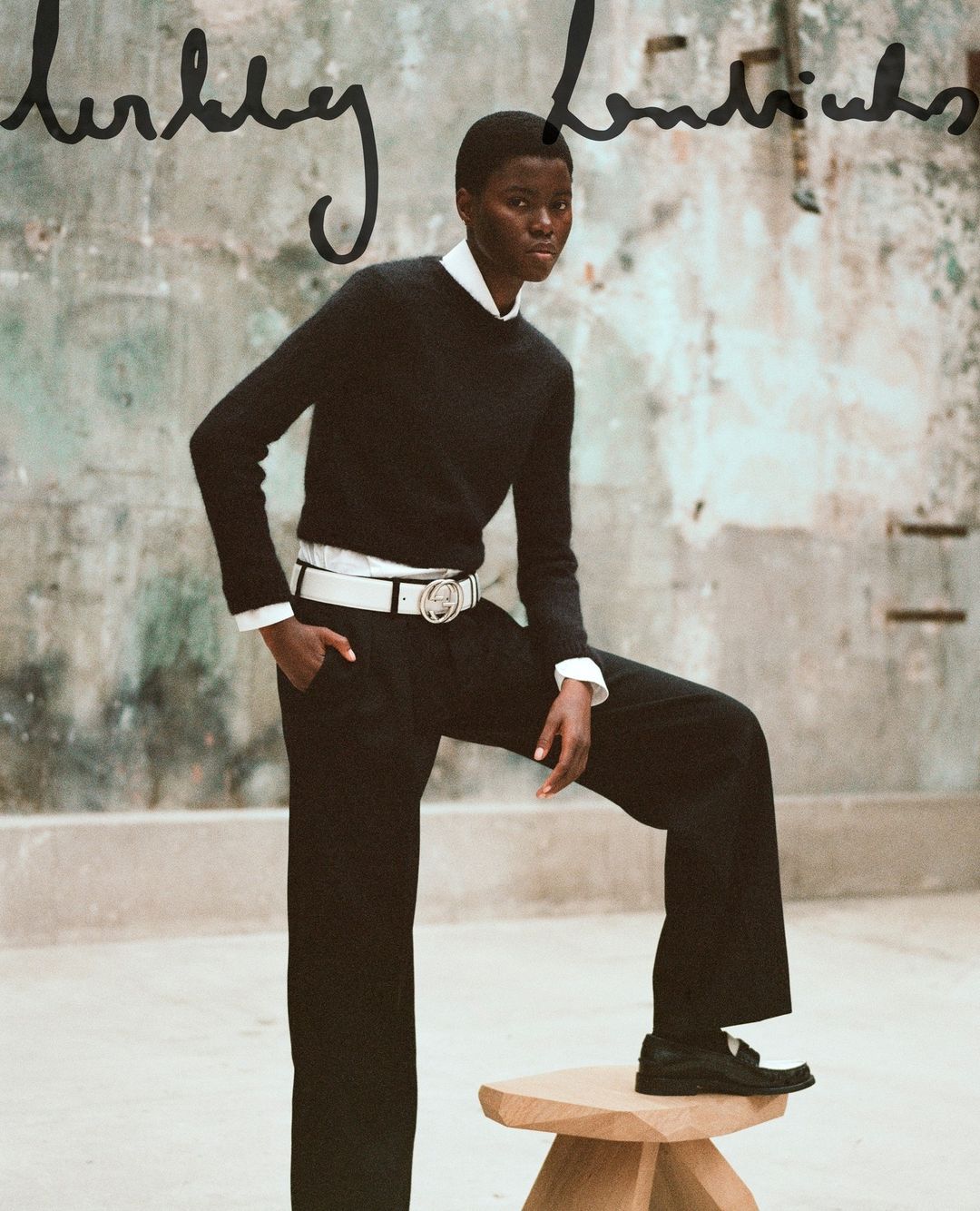 A Black female fashion model poses in black wide-leg trousers and a sweater with a white belt, indoors against an aged, neutral backdrop.