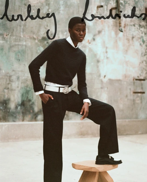 A Black female fashion model poses in black wide-leg trousers and a sweater with a white belt, indoors against an aged, neutral backdrop.