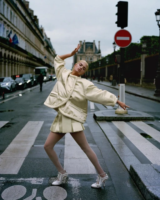 Fashionable woman in a beige jacket and skirt strikes a pose in a Paris street, with historic buildings and a traffic light in the background.