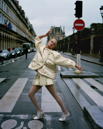Fashionable woman in a beige jacket and skirt strikes a pose in a Paris street, with historic buildings and a traffic light in the background.