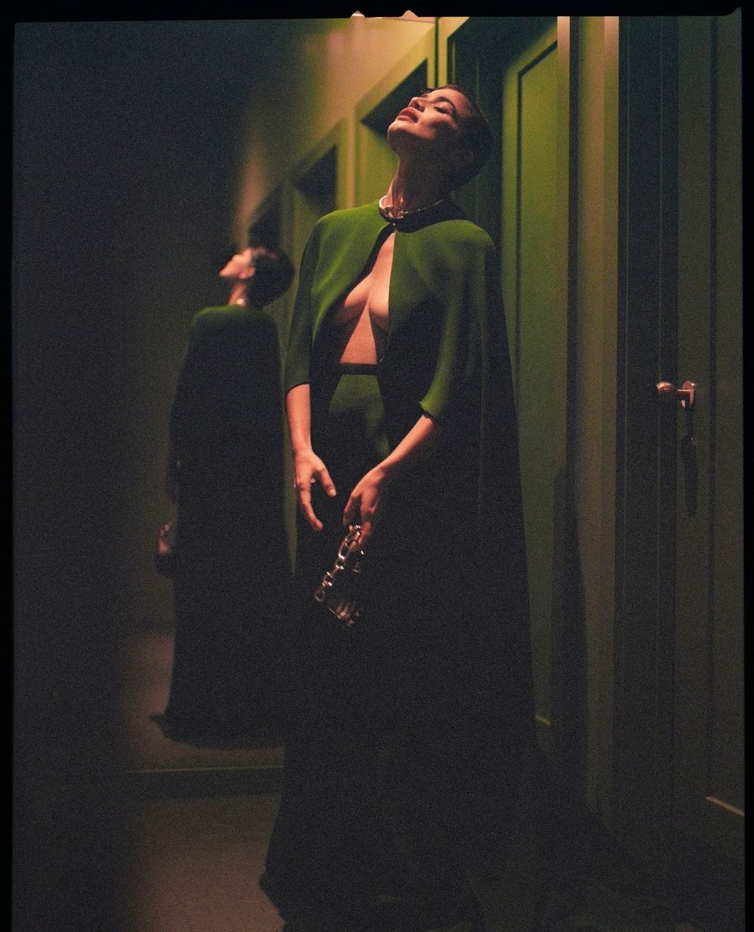 Elegant woman in green dress and cape standing in hallway with mirror reflection in green lighting; fashion, indoor, adult female.