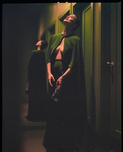 Elegant woman in green dress and cape standing in hallway with mirror reflection in green lighting; fashion, indoor, adult female.