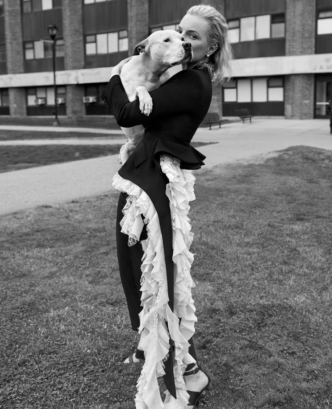 Woman in couture fashion holds a dog outside a brick building, featuring black and white tones, contrasted textures, and architectural backdrop.