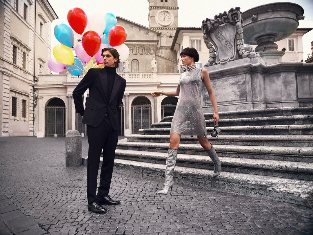 A fashionable couple is on location in an Italian town square near a fountain with colorful balloons. The woman wears a silver dress and boots.