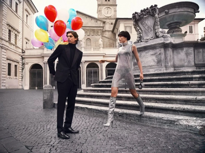A fashionable couple is on location in an Italian town square near a fountain with colorful balloons. The woman wears a silver dress and boots.