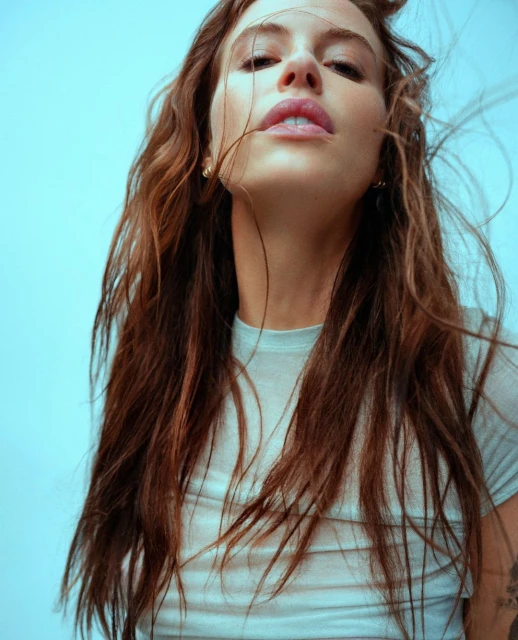 Close-up shot of an adult woman with long brunette hair, wearing a light blue top, against a light blue background.