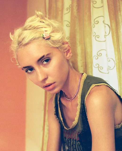 Portrait of a young adult woman with blonde hair, wearing a grey top and pink necklace, posing indoors near a patterned curtain.