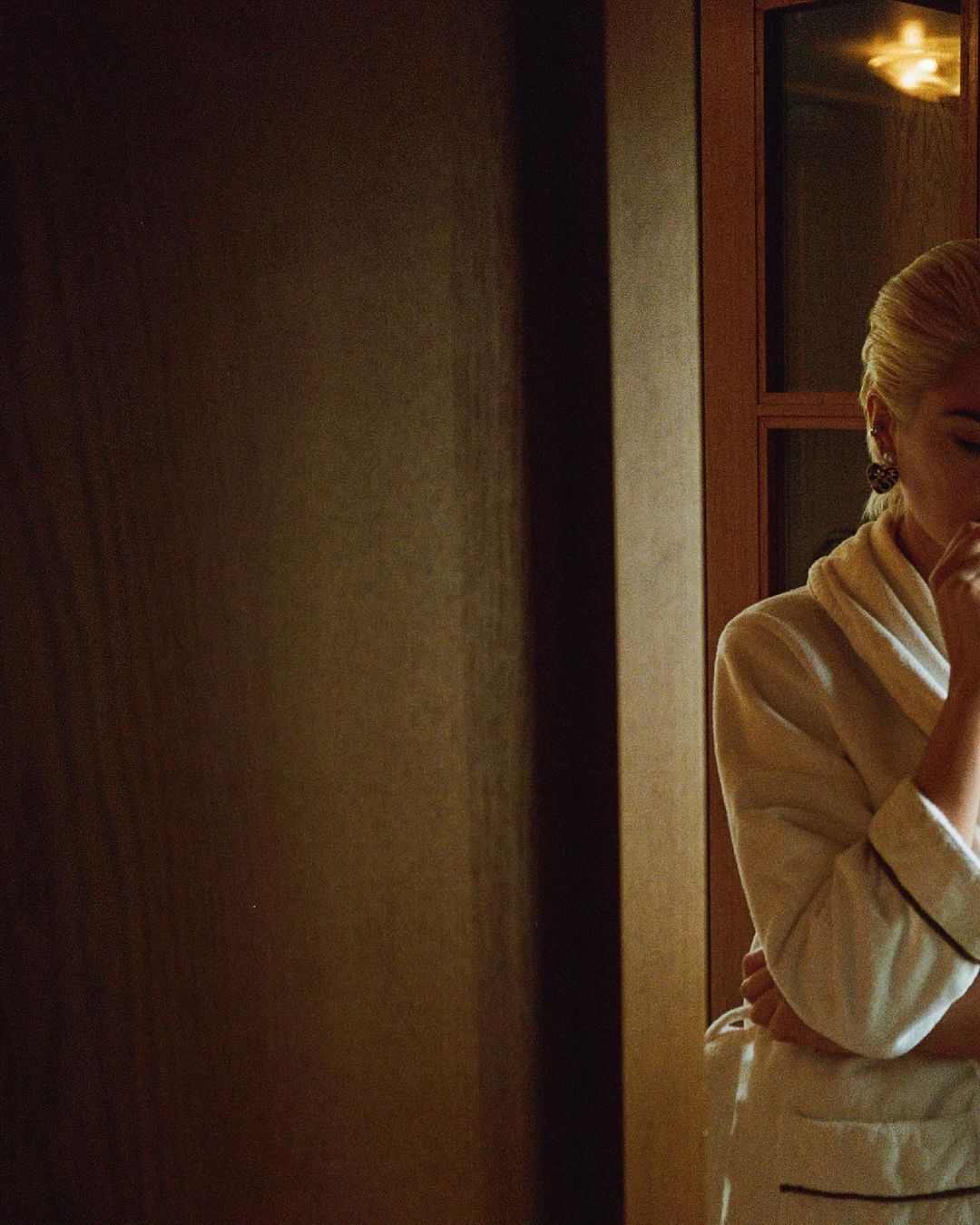 Adult woman in white robe with blonde hair stands near a dark wall and window, thoughtfully holding her chin.