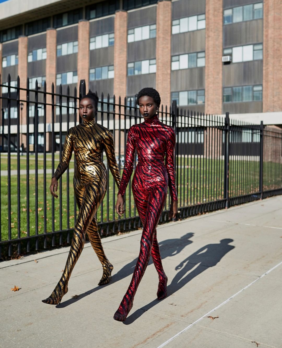 Two black female fashion models in sequined catsuits walking on a sidewalk in front of a brick building and a black metal fence.