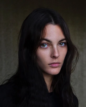 Portrait of a woman with dark hair and hazel eyes looking directly at the camera against a dark background