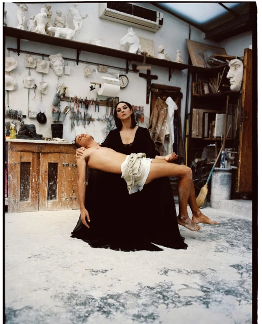 Woman in black dress supports shirtless man in indoor photo studio surrounded by art, sculptures, and a cross on the wall.
