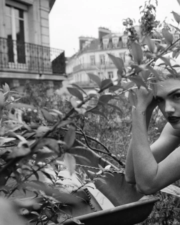 Monochrome portrait of a woman looking at camera, framed by leafy foliage and urban rooftop view.