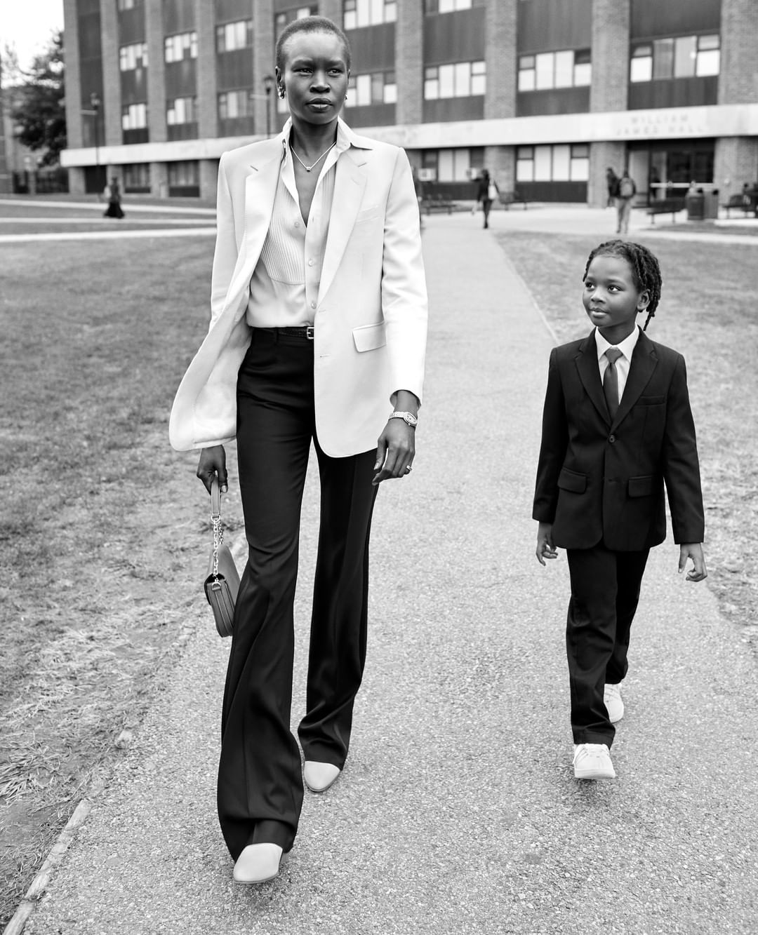 Black and white shot of an adult and a child walking on a paved path. Woman wears blazer and flared pants, child wears suit and tie.