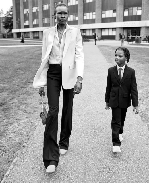 Black and white shot of an adult and a child walking on a paved path. Woman wears blazer and flared pants, child wears suit and tie.