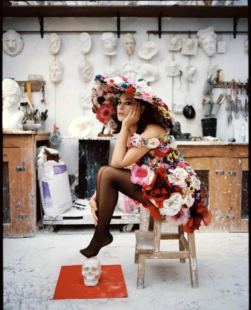 A model sits on a wooden stool in a flower-covered dress and hat, striking a pose with a skull, in a brightly lit artist's studio.
