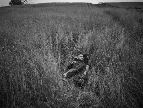 Monochrome outdoor portrait of a bearded adult man lying in tall grass in a field on a cloudy day, photographed at eye level.
