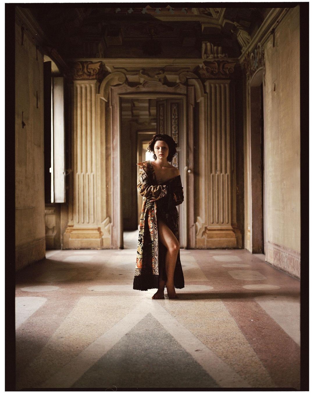 A woman stands in a long, ornate hallway wearing a patterned robe, looking at the camera with a neutral expression in editorial fashion photography.