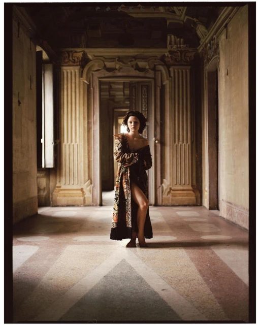 A woman stands in a long, ornate hallway wearing a patterned robe, looking at the camera with a neutral expression in editorial fashion photography.