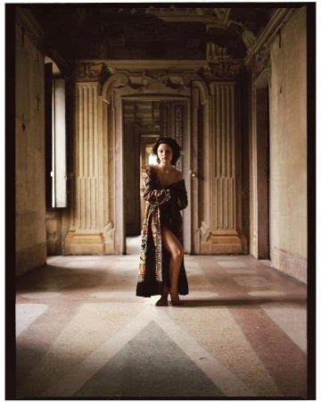 A woman stands in a long, ornate hallway wearing a patterned robe, looking at the camera with a neutral expression in editorial fashion photography.