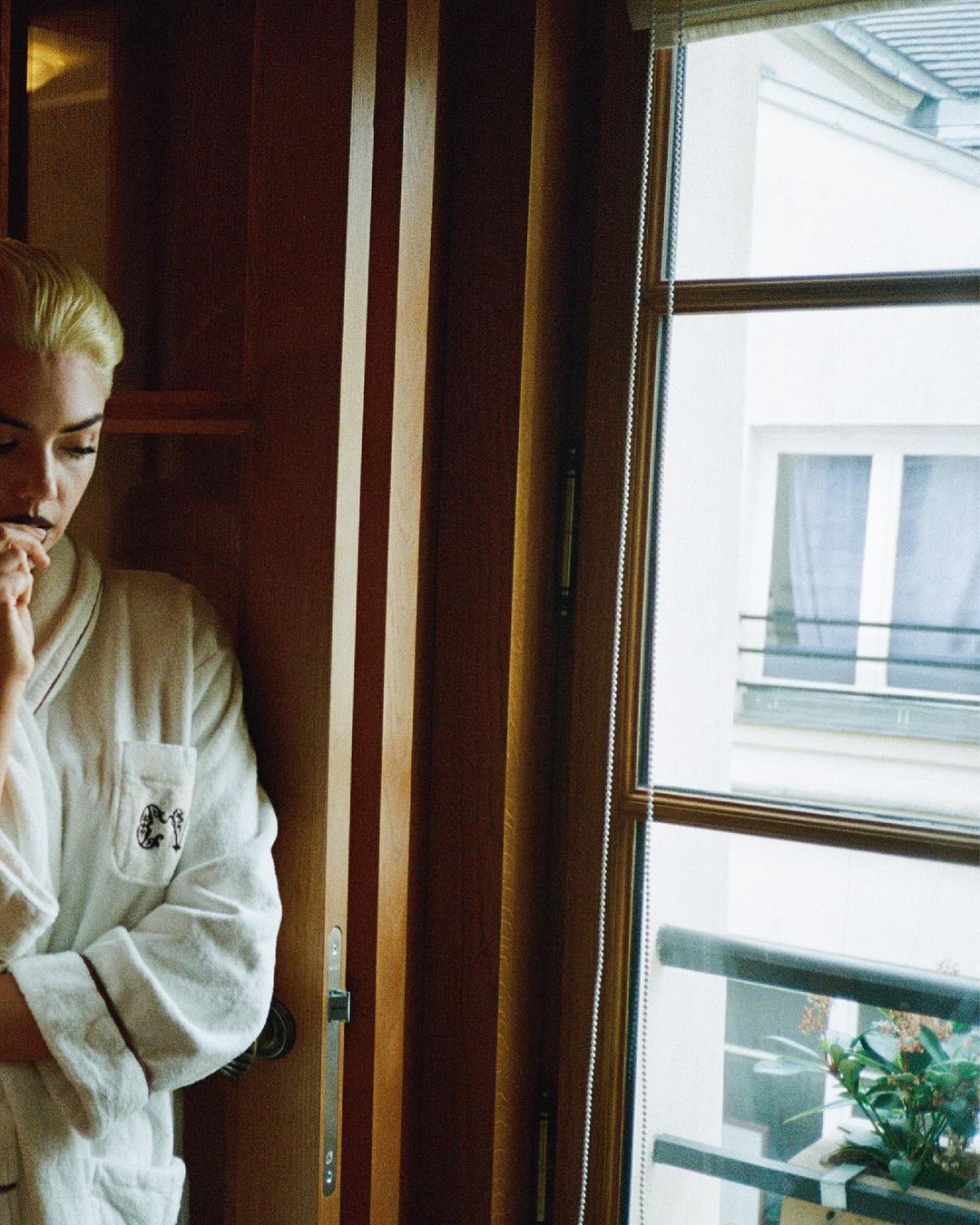 A woman with short blonde hair is partially visible wearing a white robe in a room with natural light from the window behind her.