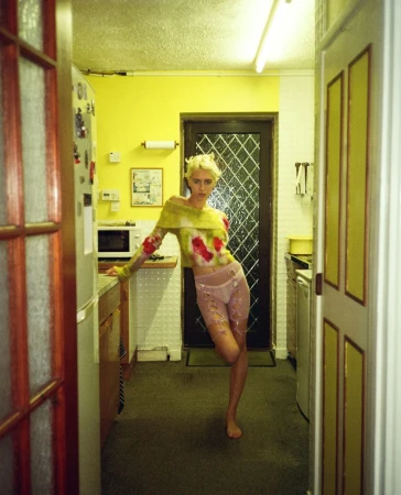 Blonde woman poses in a kitchen, wearing a colorful long-sleeve top and patterned short bottoms, leaning against a kitchen counter.