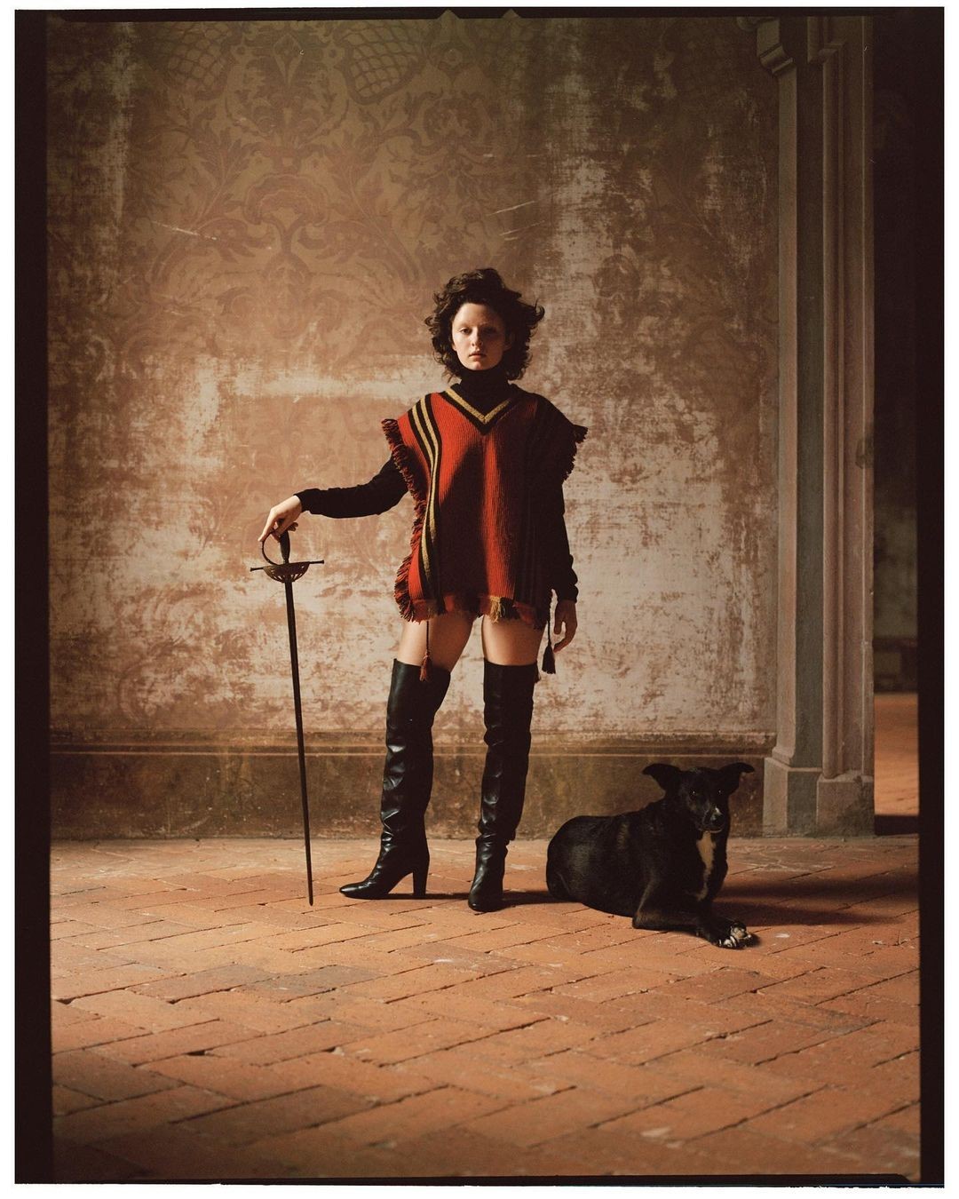 A woman in a red poncho and black thigh-high boots poses with a sword, a black dog lies on the terracotta-tiled floor nearby. Indoor scene.