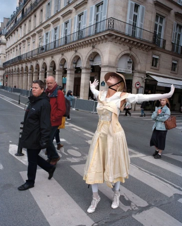 Woman in pale tiered dress and hat mid-leap on a zebra crossing with onlookers watching in a city street