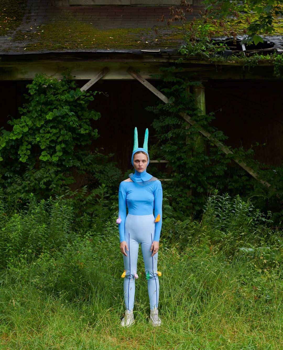 A person in a blue outfit with bunny ears stands in a grassy field in front of an old building overgrown with green vegetation.