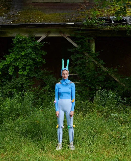 A person in a blue outfit with bunny ears stands in a grassy field in front of an old building overgrown with green vegetation.
