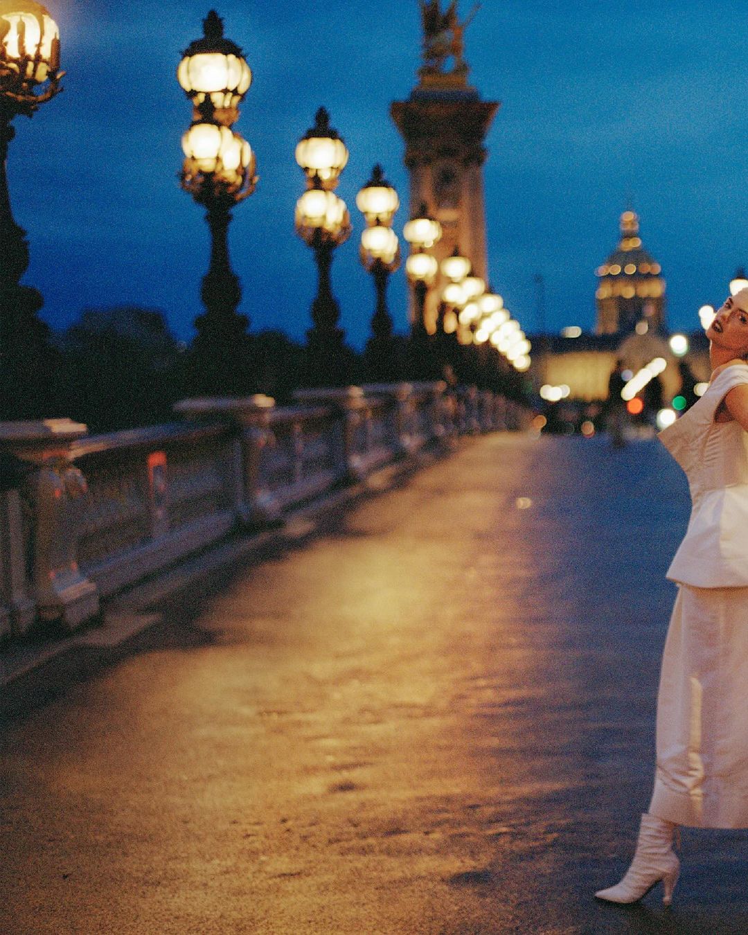 Fashionable woman in dress and boots posing on ornate bridge with streetlights and a domed building in the background at dusk.