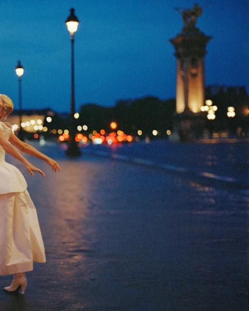 Elegant woman in a wedding dress balances on a Paris street at night near a bridge with streetlights and blurred traffic.