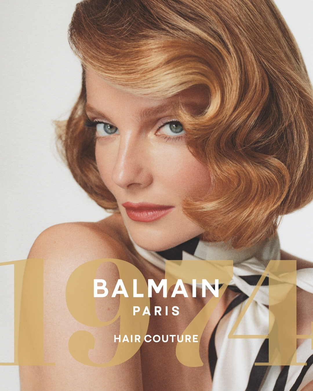 Close-up portrait of a blonde woman with vintage-styled waves, wearing a cream and black scarf, against a plain white background.