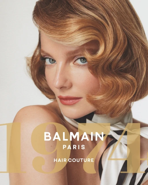Close-up portrait of a blonde woman with vintage-styled waves, wearing a cream and black scarf, against a plain white background.
