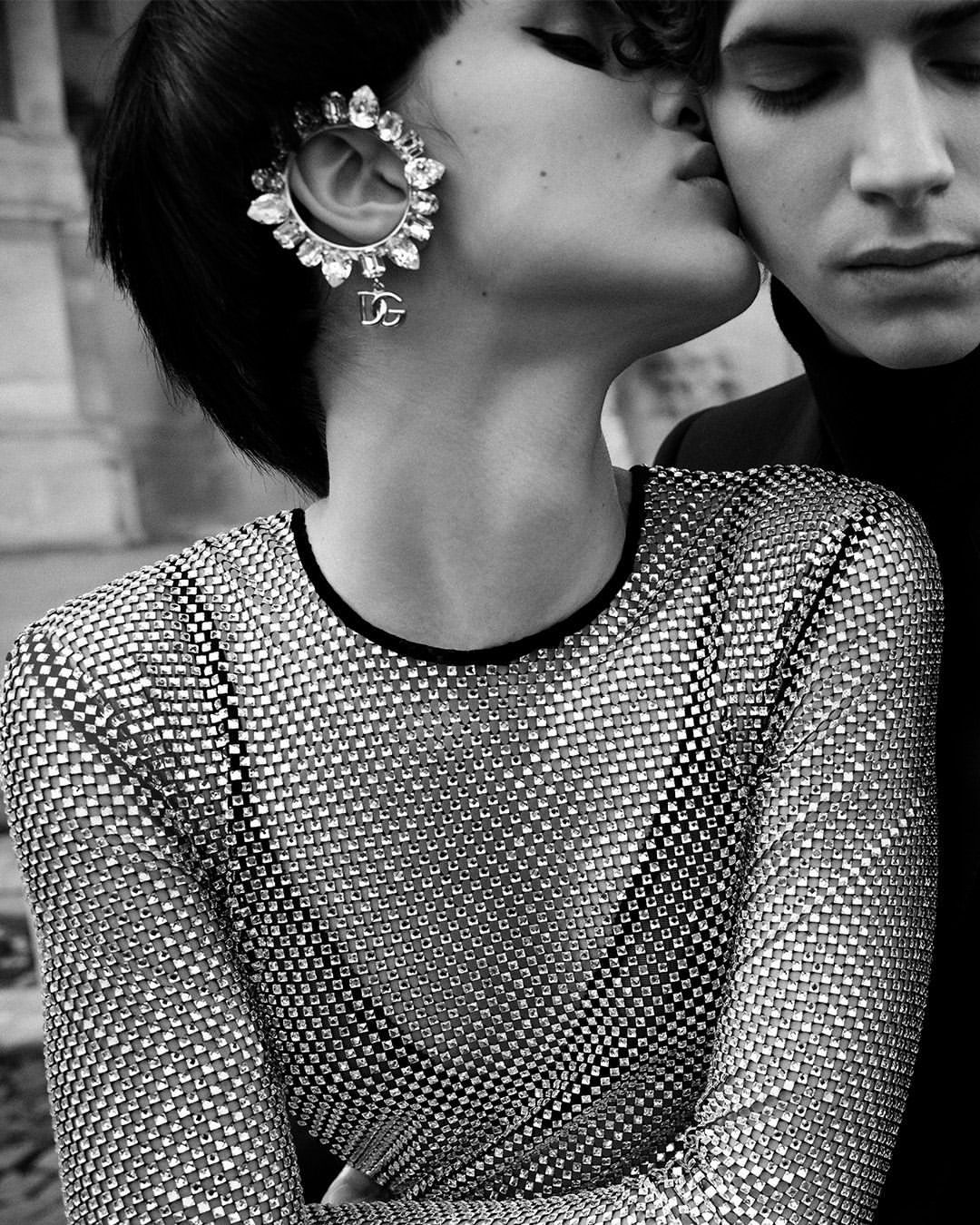 Black and white portrait of a romantic couple, featuring a woman in a jeweled top and earring, being kissed by a man in a black turtleneck.