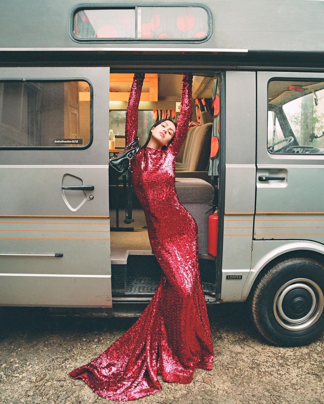 A woman in a long, red sequined dress strikes a pose with her arms raised, standing in the doorway of a gray camper van.