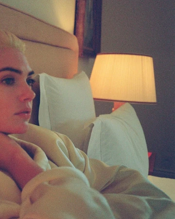 A blonde woman in a cream robe resting in a softly lit bedroom with a bedside lamp by photographer Sebastian Faena.