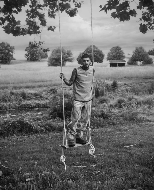 Monochrome portrait of a man standing on a rope swing in a field, wearing casual clothing, with trees and sky visible in the background.