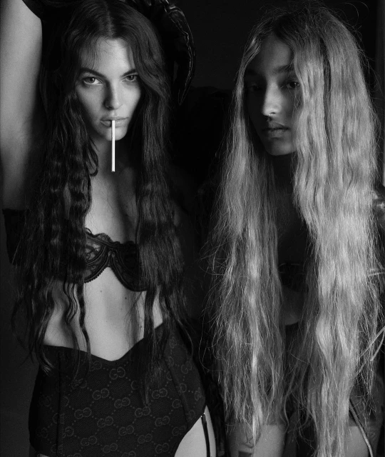 Two women with long hair pose indoors against a dark background, wearing lingerie; one holds a lollipop in her mouth, in black and white.