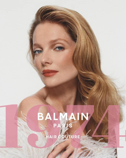 Portrait of a blonde woman with glamorous wavy hair, wearing white feathers, and a pink logo overlay for Balmain Hair Couture.