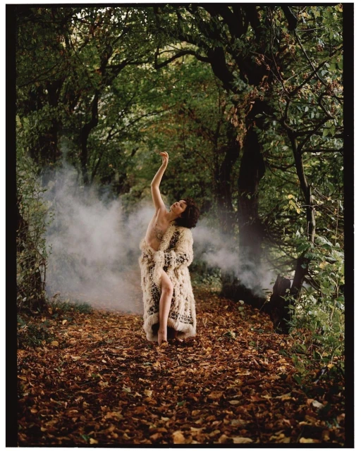 Woman in an animal print coat poses in a forest clearing filled with leaves and light smoke. Trees surround the area.