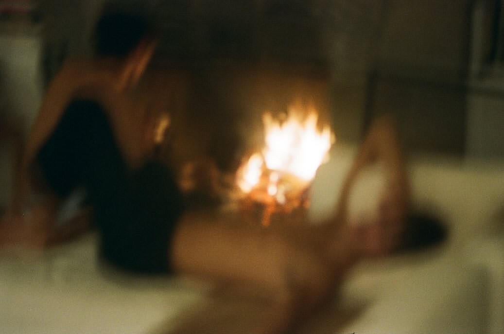 Blured indoor image of two adults, one sitting, one lying down near burning wood, with neutral colors and an out-of-focus style.